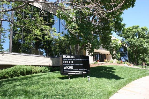 Photo of the State Higher Education Policy Center, a office building with several outward facing windows and trees around it with a sign in front with three organizations listed: NCHEMS, SHEEO, WICHE. 